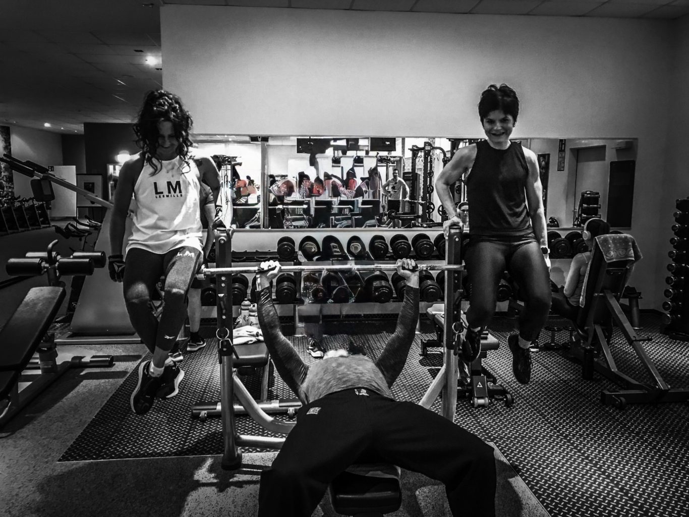 Mann drückt Langhantel im Liegen hoch und auf den Enden sitzen rechts und links je eine Frau